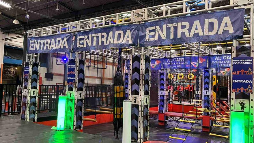 Entrada a un circuito de obstáculos tipo ninja en un parque interior, con estructuras metálicas, cuerdas y luces de colores.