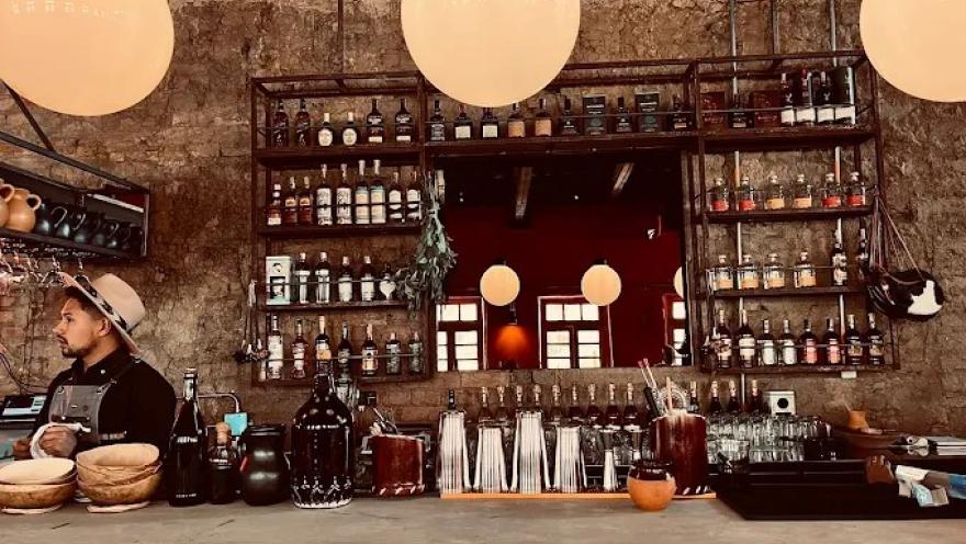 Barra de un bar con estilo rústico e industrial, con paredes de ladrillo y estanterías metálicas llenas de botellas de licor y frascos. Sobre la barra de madera se ven utensilios de coctelería, vasos y recipientes. Tres lámparas redondas cuelgan del techo, iluminando el espacio con luz cálida.