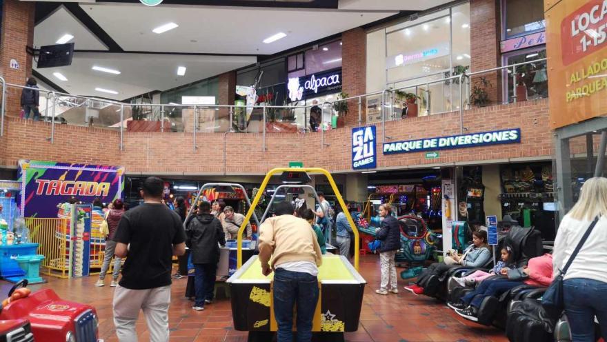  Interior de un centro comercial con zona de juegos, donde varias personas participan en máquinas arcade y mesas de juego, rodeadas de locales y señalización de entretenimiento.