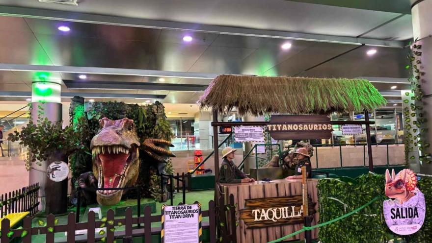  Entrada temática de dinosaurios en un espacio interior, con una gran cabeza de T-Rex, taquilla y señalización de salida