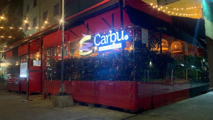 Restaurante con estructura exterior en color rojo ubicado en una esquina urbana durante la noche. El lugar tiene paredes transparentes, iluminación cálida y luces colgantes que crean un ambiente acogedor. En el interior se observan plantas decorativas y mesas ocupadas, mientras en la fachada resalta un letrero iluminado con el nombre “Carbu”.