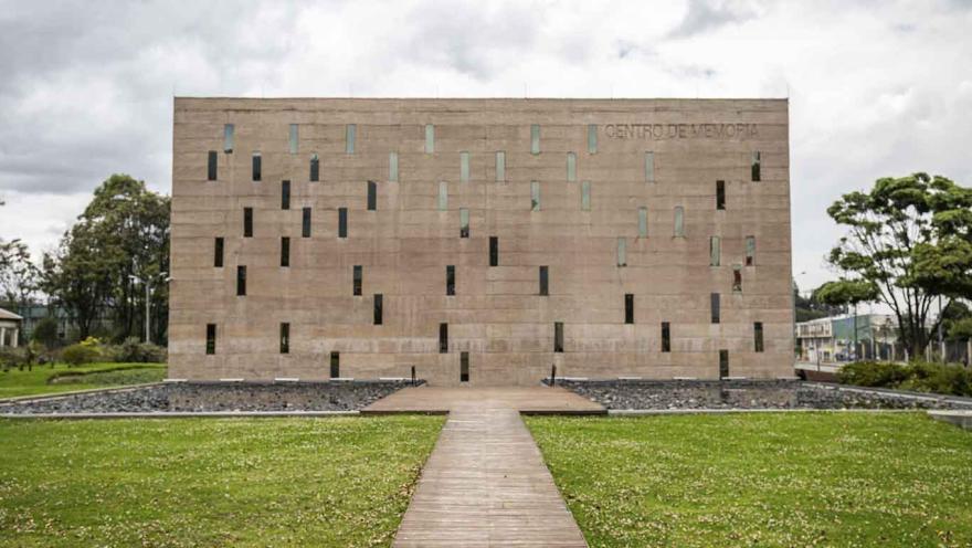 La imagen muestra un edificio de arquitectura moderna y minimalista, con una fachada de piedra clara perforada por ventanas verticales irregulares. Un sendero recto conduce hacia la entrada, atravesando una zona verde que rodea la estructura.