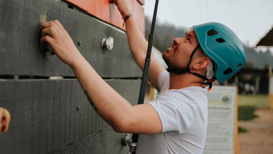 Persona con casco y arnés escala un muro de madera con agarres, en una actividad de aventura al aire libre.