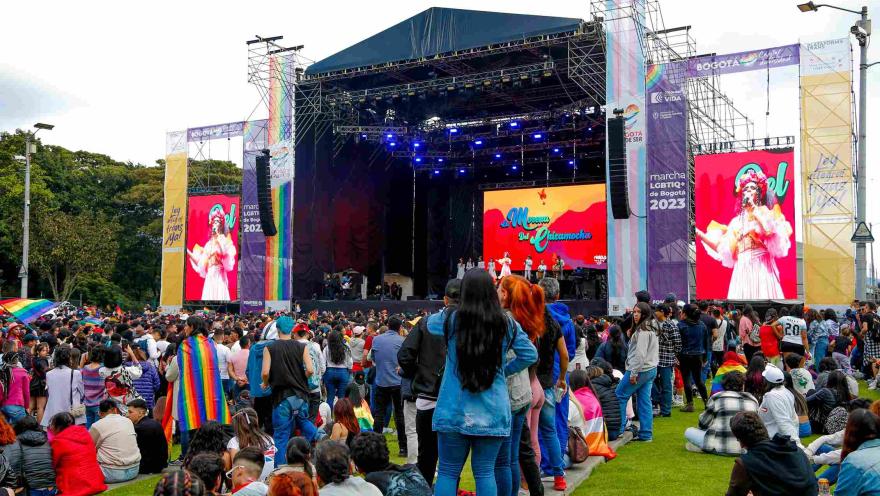 Multitud de personas reunidas frente a un gran escenario al aire libre durante un evento del orgullo LGBTI+ en Bogotá; en las pantallas se ve una artista cantando, mientras asistentes llevan banderas arcoíris y observan el espectáculo sobre el césped.