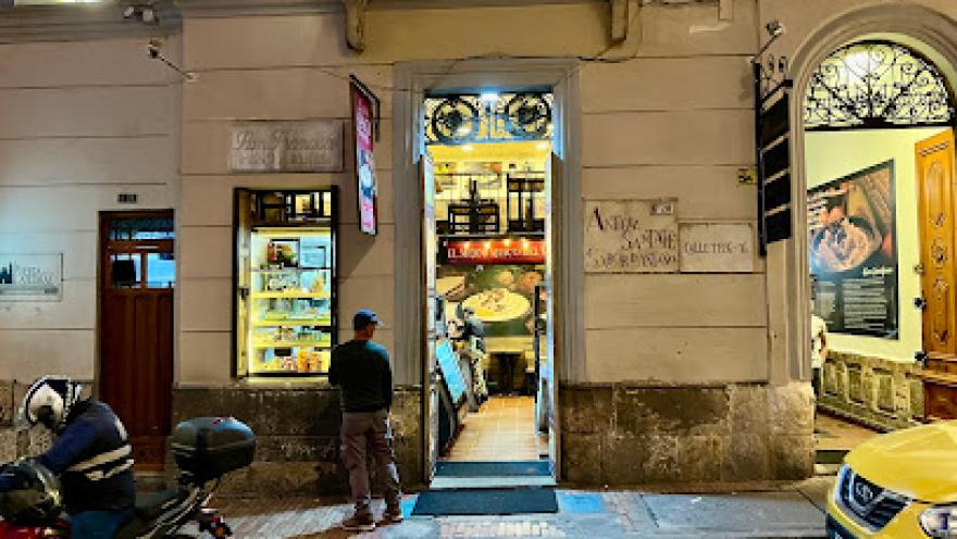 Fachada nocturna de un restaurante tradicional en una calle del centro histórico. La puerta abierta deja ver el interior iluminado con mesas dispuestas para los comensales, mientras una vitrina exhibe productos o alimentos. Afuera, una persona observa el lugar y a un costado hay motocicletas estacionadas y un taxi amarillo, típico de la ciudad.