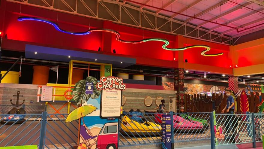Atracción de carros chocones en un parque de entretenimiento interior, con luces de neón y vehículos de colores dentro de una pista cerrada.