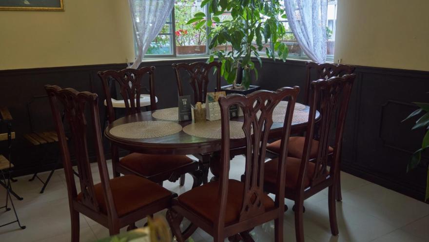 La imagen muestra un comedor de estilo clásico, con una mesa redonda de madera rodeada por sillas talladas en tonos oscuros. La luz natural entra por una ventana con cortinas ligeras, iluminando una planta ubicada en el centro.