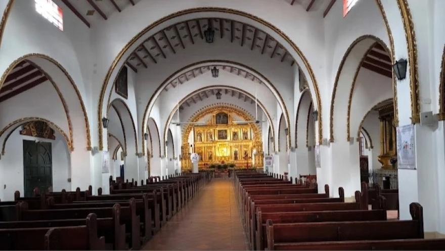 La imagen muestra el interior de una iglesia de estilo colonial, caracterizada por su amplitud y simetría. Un largo pasillo central de piso en tonos cálidos conduce la mirada hacia el altar mayor, mientras a ambos lados se alinean filas de bancas de madera oscura, ordenadas y silenciosas, que refuerzan la sensación de recogimiento y solemnidad.