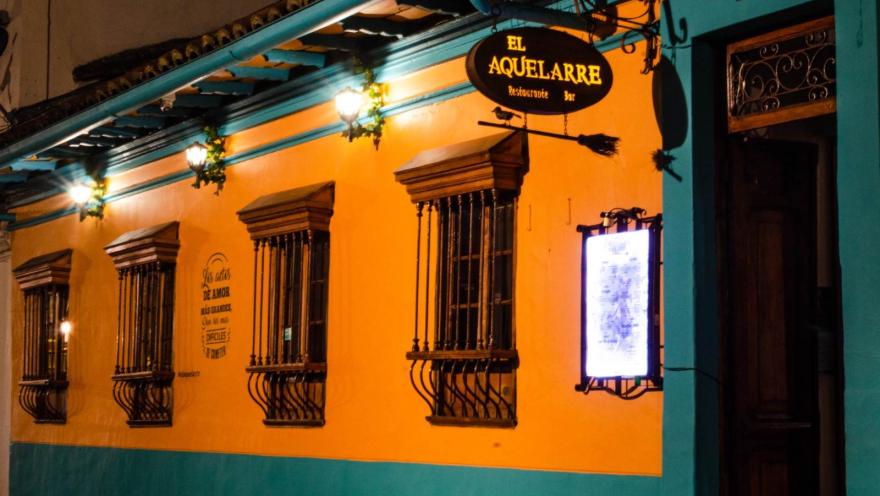 Fachada nocturna de un restaurante-bar de estilo colonial, pintado en tonos amarillo y turquesa. En la parte superior cuelga un letrero iluminado que dice “El Aquelarre”, acompañado de una escoba decorativa. La pared tiene varias ventanas con rejas de hierro y faroles encendidos que aportan una luz cálida.