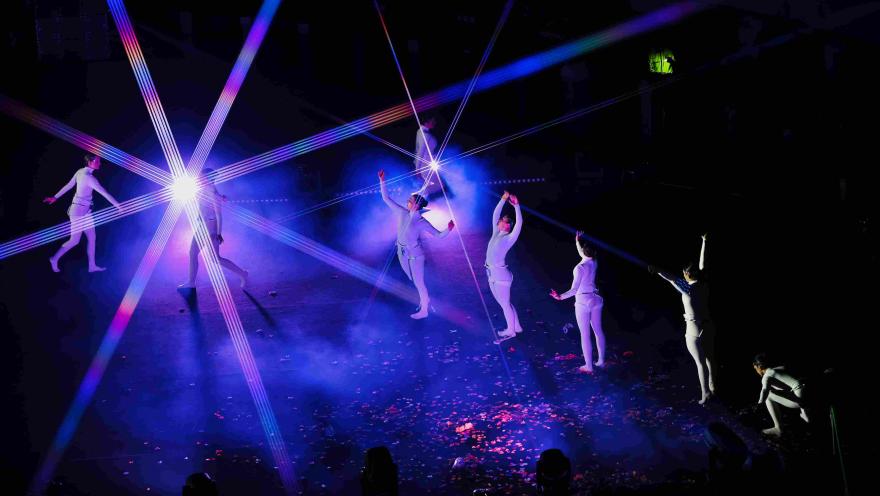  Escena teatral con varios bailarines vestidos de blanco sobre un escenario oscuro, iluminados por luces de colores y rayos láser que cruzan el espacio. Algunos performers están de pie y otros en movimiento, rodeados de humo y confeti en el suelo.