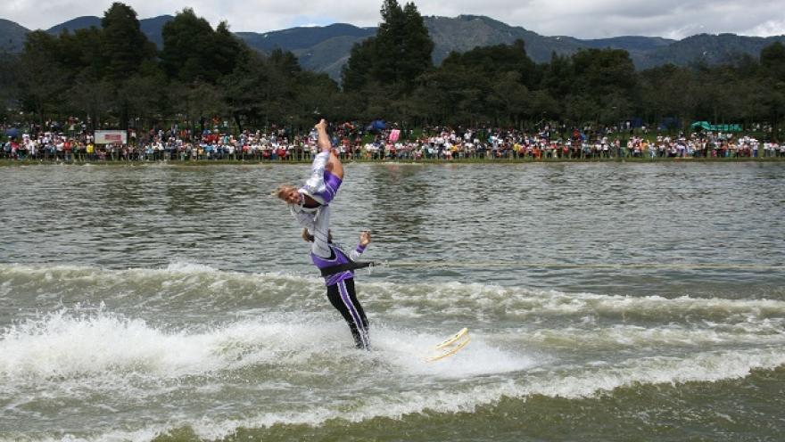 Persona practicando esquí acuático en un lago, realizando una acrobacia frente a espectadores en la orilla.