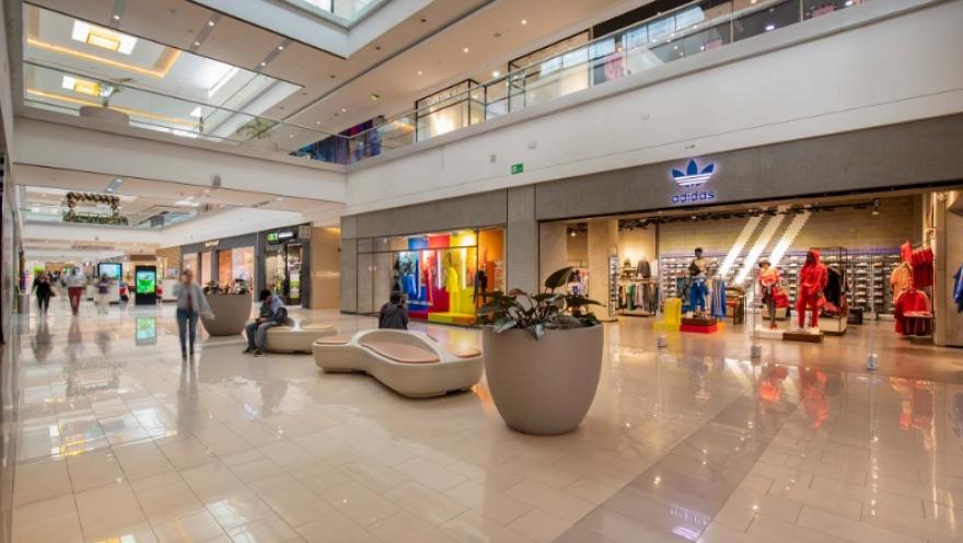  La imagen muestra el interior de un centro comercial moderno y elegante, con amplios pasillos iluminados por luz natural que entra desde el techo. A un lado se observa una tienda de ropa con vitrinas coloridas y maniquíes, mientras que en el centro hay mobiliario de descanso y una gran maceta con plantas.