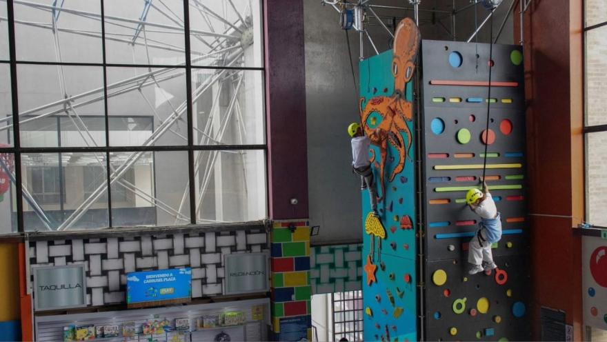 Dos niños con casco escalan un muro de escalada interior con paneles coloridos y figuras decorativas.