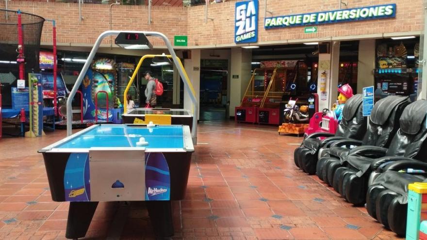 Zona de juegos en un centro comercial con mesa de air hockey, máquinas arcade y sillas de masaje, bajo un letrero de parque de diversiones.