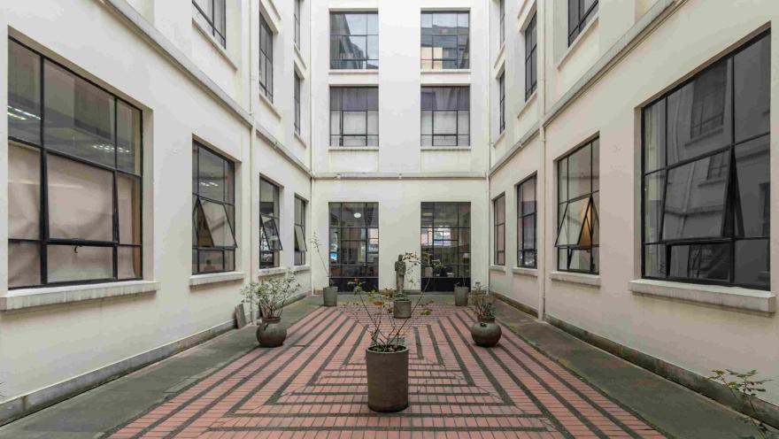 La imagen muestra un patio interior de estilo sobrio y ordenado, rodeado por paredes claras y grandes ventanas. En el centro, varias macetas con plantas se distribuyen sobre un piso de ladrillo dispuesto en patrones geométricos, creando un ambiente tranquilo y equilibrado.