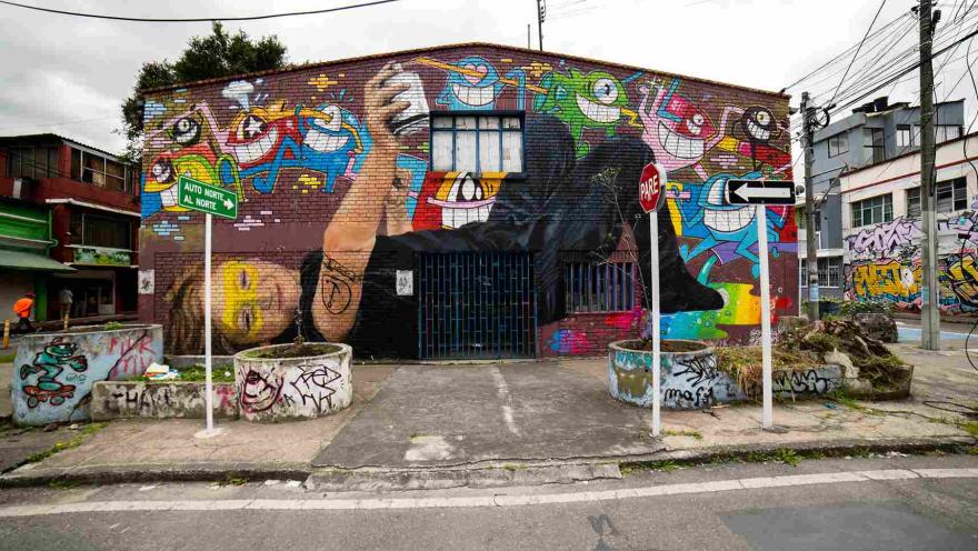 Facade of a building with a large, colorful mural that blends faces, abstract figures, and graffiti; traffic signs and an urban street are visible in front.