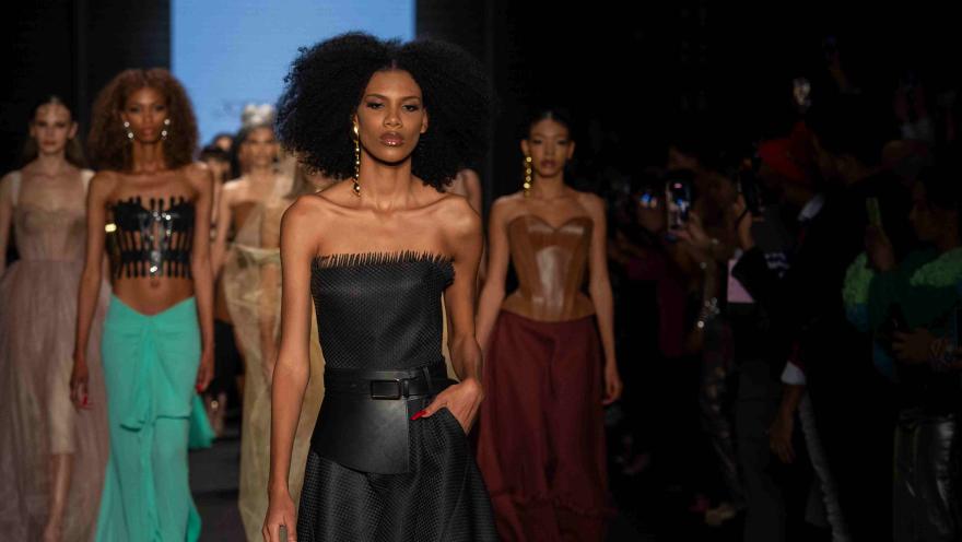Modelo con vestido negro desfila en pasarela, seguida por otras modelos con atuendos elegantes ante el público.