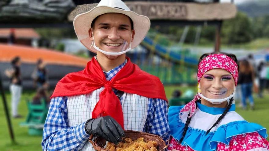 Dos personas con trajes tradicionales sonríen y muestran una canasta con comida típica en un parque al aire libre.