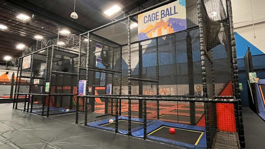 Área de juego “cage ball” en un parque interior, con canchas enrejadas, trampolines y pelotas dentro de una estructura cerrada.