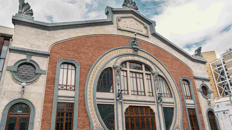La imagen muestra la fachada de un edificio histórico con detalles ornamentales y estilo clásico, donde destacan los arcos decorativos y el trabajo en piedra y ladrillo. En el centro, una gran entrada enmarcada por formas curvas y elementos artísticos resalta su carácter cultural.