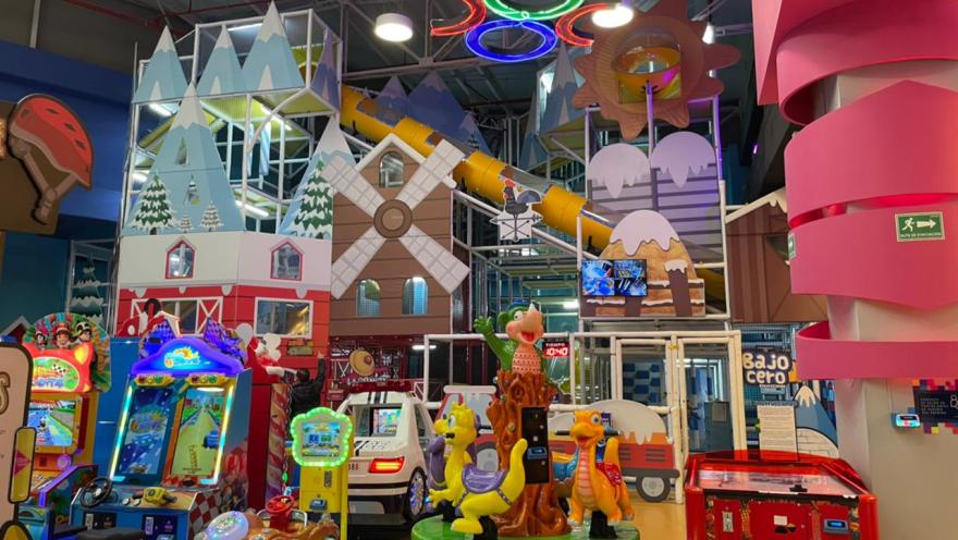Área de juegos infantiles en un parque interior con máquinas arcade, figuras coloridas y una gran estructura de juegos con toboganes y decoraciones temáticas.