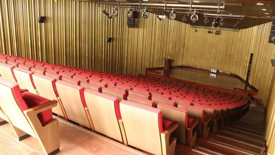 La imagen muestra un auditorio amplio y ordenado, con filas de asientos en tonos rojos dispuestos en forma descendente hacia el escenario. Al frente se observa una tarima de madera con iluminación profesional en el techo, rodeada por paredes con paneles verticales que aportan calidez y acústica al espacio.