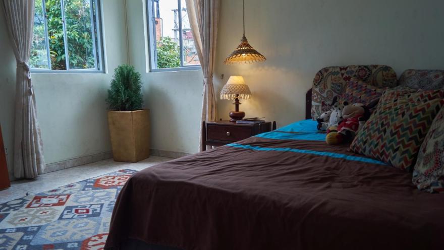 La imagen muestra una habitación acogedora y bien iluminada, con una cama vestida en tonos cálidos y cojines coloridos. La luz natural entra por dos ventanas con cortinas claras, complementada por una lámpara de mesa que aporta calidez.