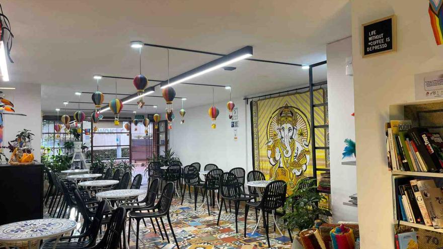 A spacious, bright café with round tables and black chairs, colorful tile floors, and balloon-shaped pendant lights. In the background, a mural of Ganesha in shades of yellow and a plant-filled area; to the right, a bookshelf and a sign that reads “Life without coffee is depresso.”