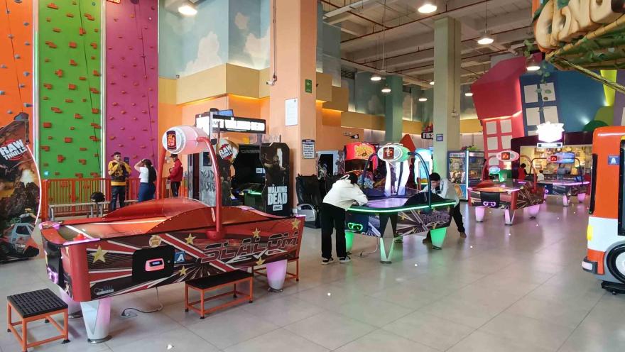Área de juegos arcade en interior con mesas de air hockey, máquinas recreativas y un muro de escalada al fondo.