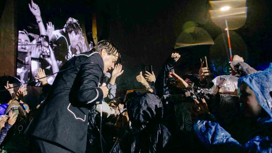 Cantante bajo la lluvia se acerca al público, que levanta las manos y celulares mientras disfrutan el concierto.