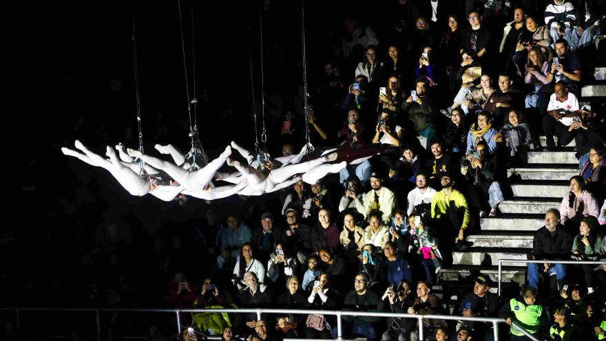  Espectáculo en vivo con acróbatas vestidos de blanco suspendidos horizontalmente en el aire, mientras un público numeroso observa desde gradas, muchos grabando con sus celulares en un ambiente nocturno.