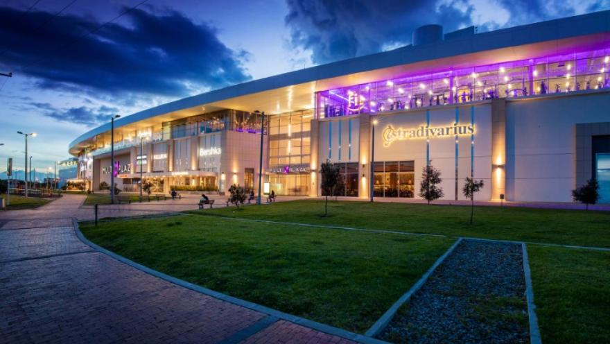 La imagen muestra un centro comercial moderno al atardecer, iluminado con luces cálidas y tonos violetas que resaltan su arquitectura contemporánea. La fachada curva alberga tiendas reconocidas y amplios ventanales, mientras al frente se extiende una zona verde con senderos peatonales. El cielo azul oscuro con nubes añade un ambiente tranquilo y atractivo a la escena.