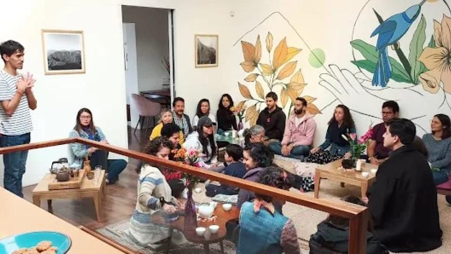 Interior of a cozy space where a group of people is participating in a guided activity. On the left, a man is standing, speaking and gesturing to the group. The participants are seated on the floor and on cushions around low tables with teapots, cups, and natural elements, suggesting an experience centered on herbal teas or wellness.