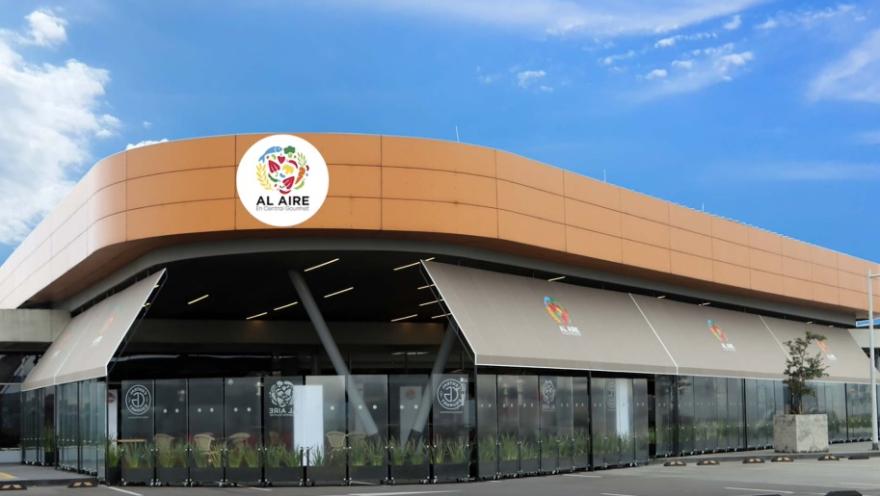  La imagen muestra un moderno espacio comercial con un diseño arquitectónico elegante y contemporáneo, identificado por el letrero “Al Aire” en su parte superior. La estructura se distingue por su forma curva y envolvente, revestida en un tono cálido similar al cobre, que contrasta armoniosamente con el cielo azul y las nubes dispersas del fondo.