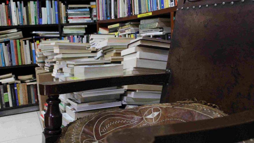 Una serie de libros arrumados detrás de una silla de madera. Al fondo hay una estantería en madera repleta de libros.