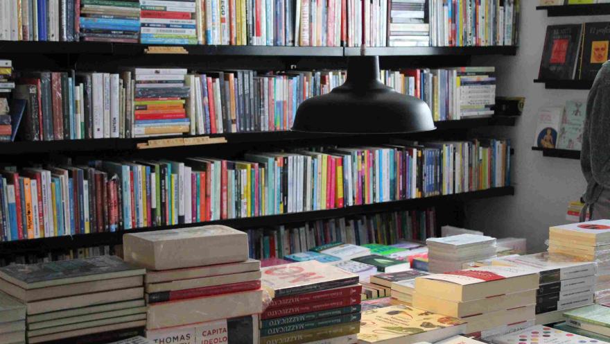 Mesa con pilas de libros bajo una lámpara negra, frente a estanterías llenas de títulos en una librería.