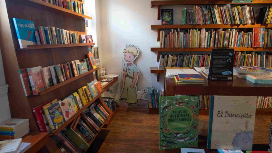 Children's section of a bookstore with wooden shelves full of illustrated books, a display table, and a figurine of The Little Prince in the background.