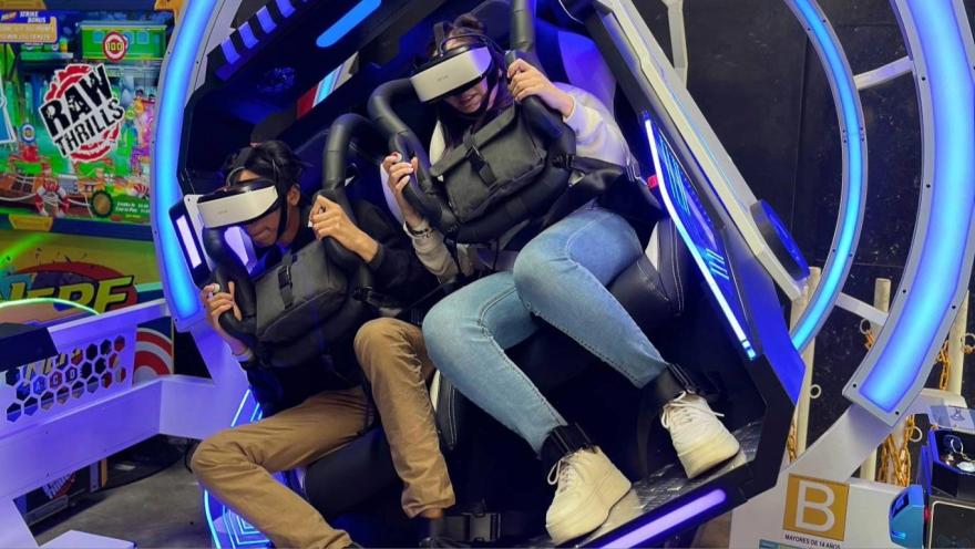 Two people use virtual reality headsets in an interactive attraction, secured to seats with harnesses inside a lighted machine.