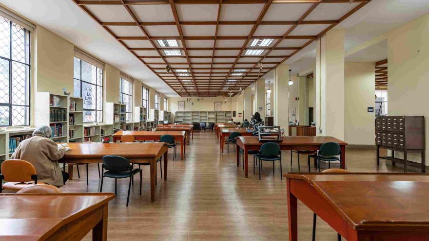La imagen muestra una sala de lectura amplia y silenciosa, con mesas de madera alineadas y estanterías llenas de libros a lo largo de las paredes. La luz natural entra por grandes ventanas, creando un ambiente tranquilo donde algunas personas leen o estudian.