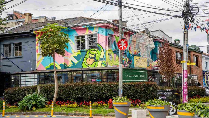 Corner of a neighborhood with a house covered by a colorful, urban-style mural; a café operates on the ground floor, surrounded by plants, cables, and traffic signs.