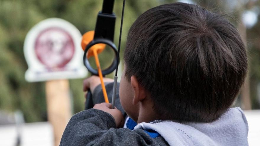 Niño practica tiro con arco apuntando hacia una diana en una actividad al aire libre.