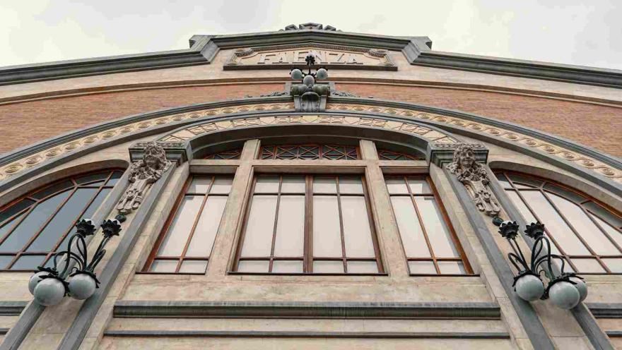 La imagen muestra un detalle de la fachada de un edificio histórico, donde destacan un gran arco decorativo y ventanales enmarcados por elementos ornamentales. Esculturas y lámparas clásicas refuerzan su estilo artístico, mientras el ángulo en contrapicado resalta la altura y elegancia de la construcción. 
