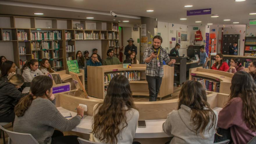  Grupo de personas participa en una actividad en biblioteca, escuchando a un facilitador en el centro del espacio.