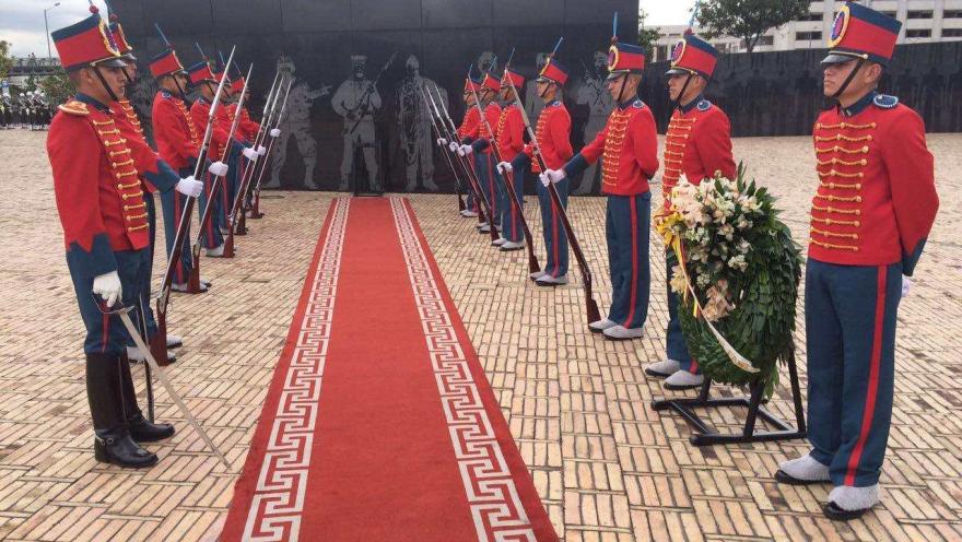  Guardia de honor con uniformes rojos custodia un monumento, junto a una ofrenda floral y una alfombra roja.