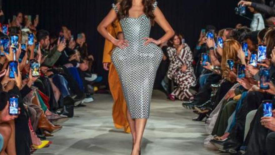  Modelo desfila en pasarela con vestido ajustado a cuadros, mientras el público toma fotos a ambos lados.