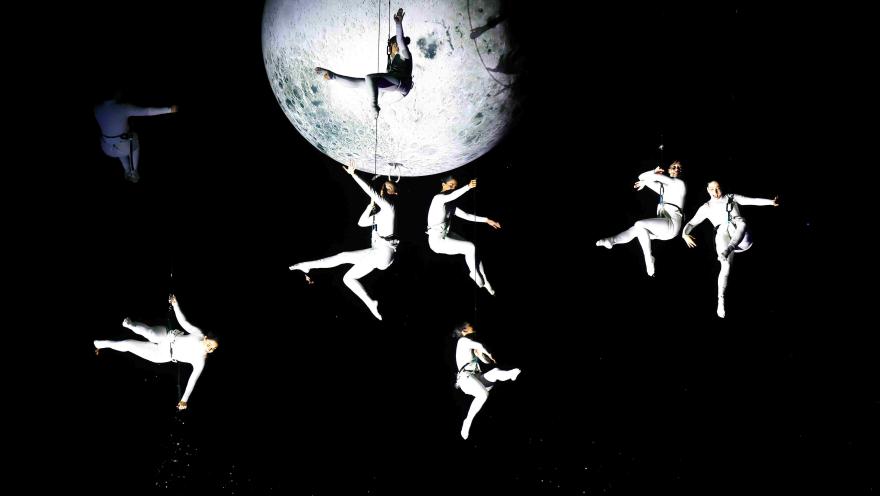  Espectáculo aéreo con varios acróbatas vestidos de blanco suspendidos en el aire alrededor de una gran luna iluminada, creando una escena poética en un fondo oscuro.