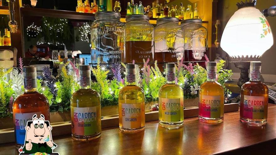 Barra de un bar iluminada con luz cálida, donde se exhiben varias botellas de ginebra alineadas en primer plano, cada una con etiquetas de diferentes sabores como cítricos y flores. Detrás, hay frascos grandes con infusiones y una repisa con más botellas de licor.