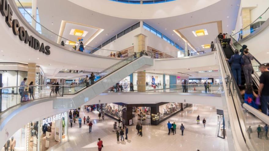 La imagen muestra el interior de un centro comercial de varios niveles, con una arquitectura amplia y luminosa. Escaleras eléctricas conectan los pisos, mientras personas transitan entre tiendas y zonas de comida. La iluminación cálida y los espacios abiertos generan una sensación de dinamismo y modernidad.