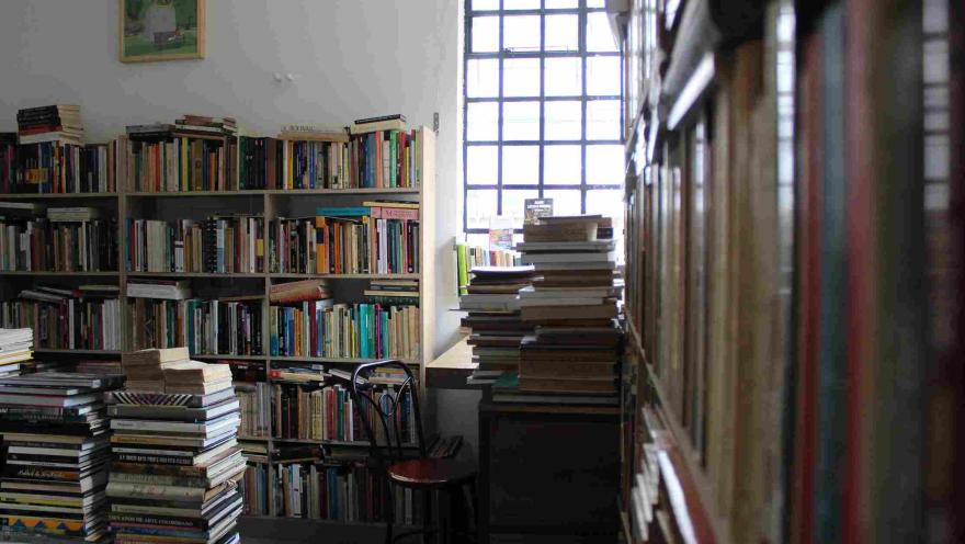 Interior de librería con estanterías llenas y pilas de libros junto a una ventana.