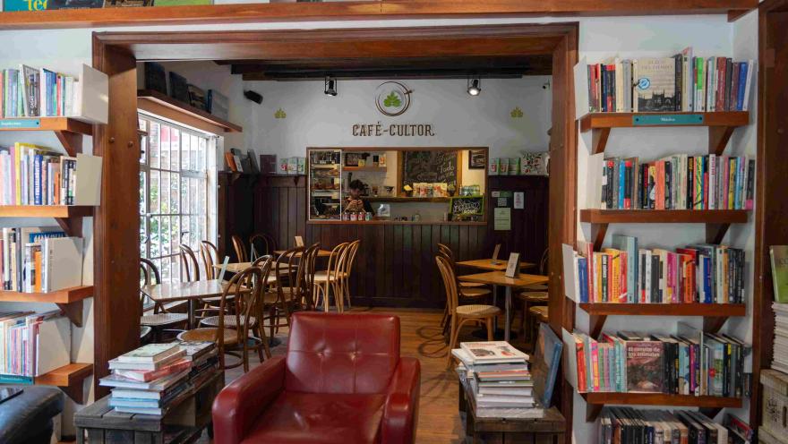 Interior of a bookstore with a café, surrounded by bookshelves, wooden tables, and a counter in the back under the sign "Café-Cultor."